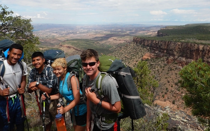 Four students wearing backpacks smile at the camera. They are high above a southwestern landscape.